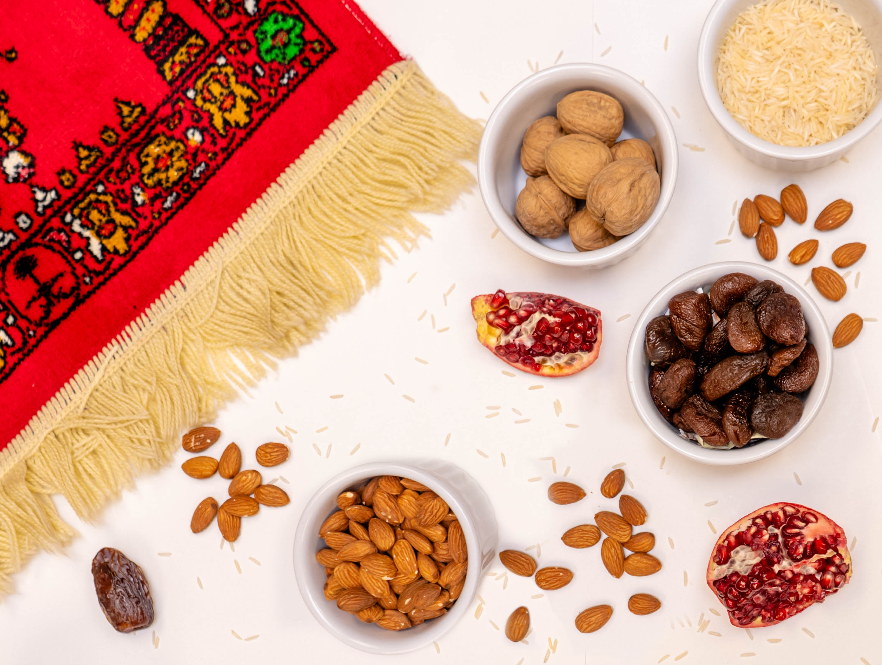 Prayer rug, pomegranate and bowls of dates, almonds, rice and walnuts on a white background.