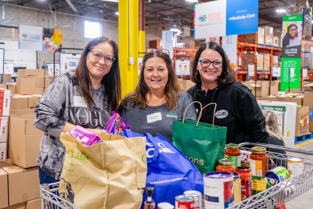 Three volunteers standing behind a cart full of shelf-stable food donations in Food Banks Mississauga's warehouse.